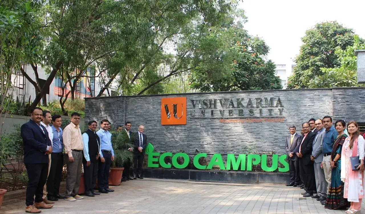 Vishwakarma University (VU) Pune Eco Campus entrance &mdash; stone signage wall with the orange VU shield and Vishwakarma University name, ECO CAMPUS in green at the base, leafy trees overhead and faculty in formal dress at the front &middot; Kondhwa Budruk green campus